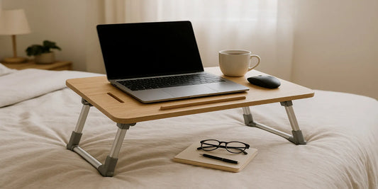 Adjustable foldable bed laptop desk with laptop, mug and mouse on beige bedding; notebook and glasses nearby in soft daylight.