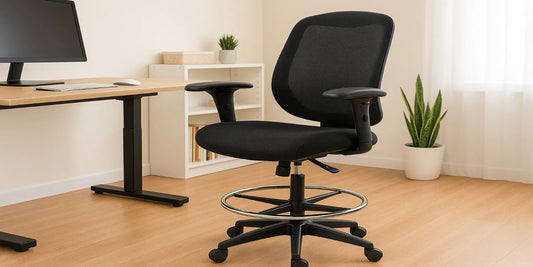 A modern ergonomic drafting chair with mesh back and foot ring placed next to a standing desk in a bright, minimalist home office setting.